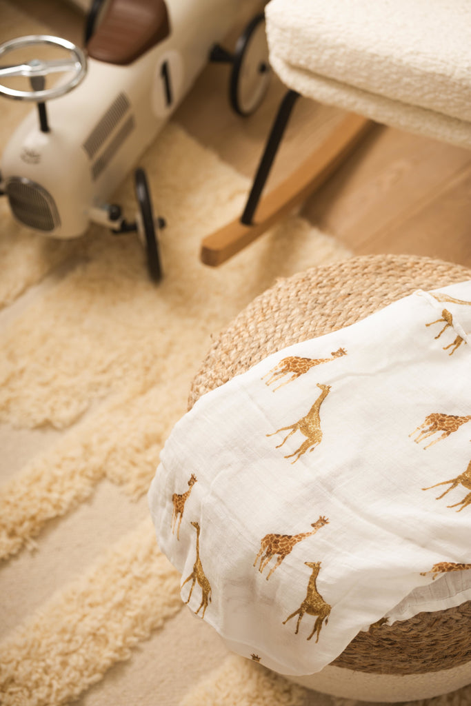 image of a watercolour giraffe printed muslin swaddle layed on a natural and white pouf