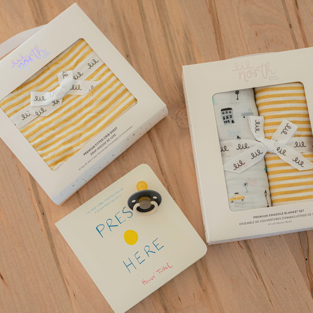 Image of a baby gift bundle. Products include a 2 pack set of swaddles in ochre stripe and a grey city cars print. the crib sheet is an ochre stripe and a book sits beside it.